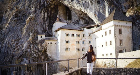 A traveler woman stands back to the camera and enjys view of Predjama Castle (Predjamski grad), a Renaissance castle built within a cave mouth in the historical region of Inner Carniola, Slovenia