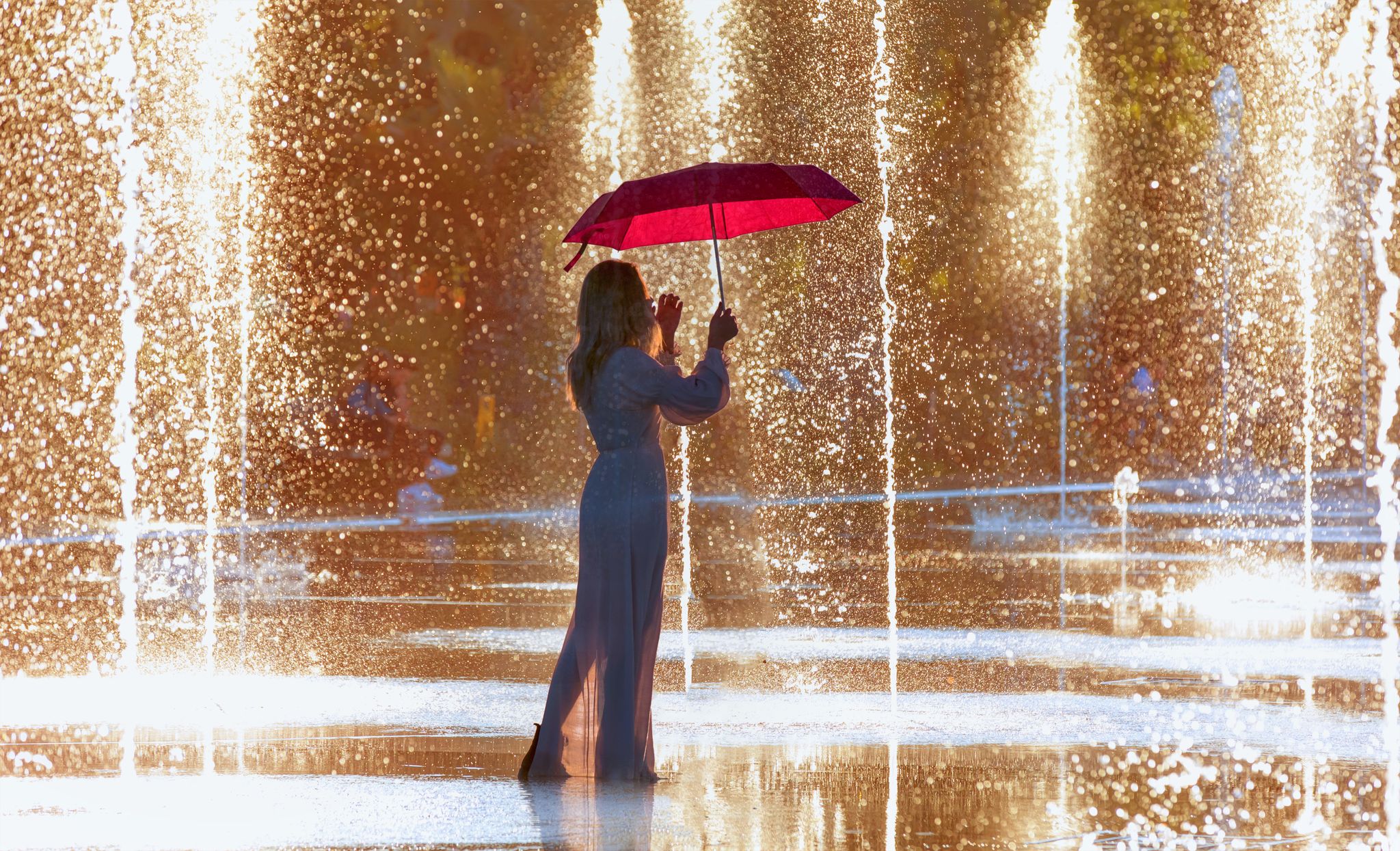 photo of young girl in white dress holding red umbrella and walking on the fountain at Picturesque Park Paillon Promenade in Nice, France.