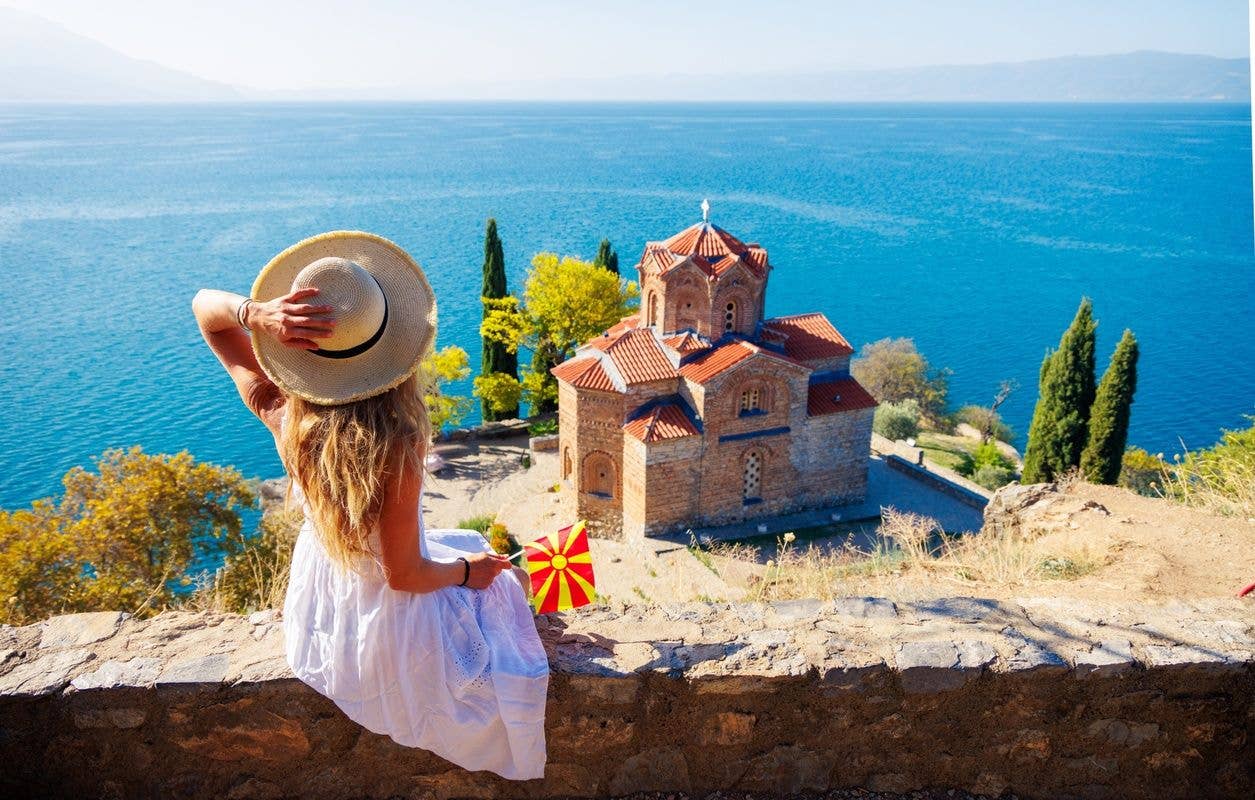 A woman in a white dress and hat sits on a stone wall, admiring the Church of St. John at Kaneo in Ohrid, North Macedonia..jpg