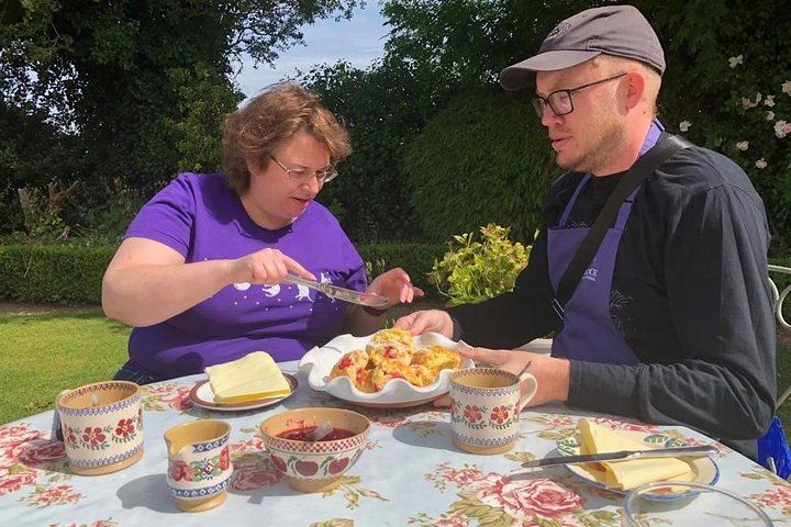 A man and a woman enjoy a meal outdoors at a floral table, with tea and pastries in a green garden setting..jpg
