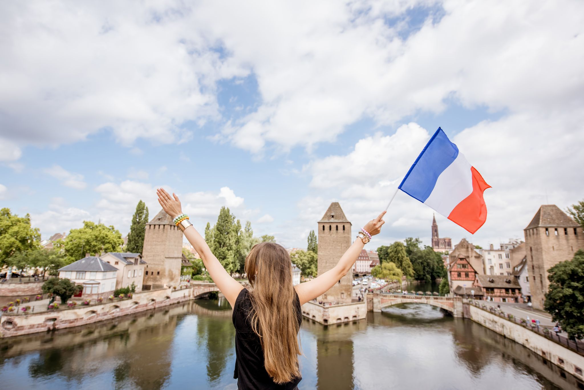 photo of young woman tourist sitting on Barrage Vauban with French flag on the beautiful landscape background with river and towers in the old town of Strasbourg city, France.