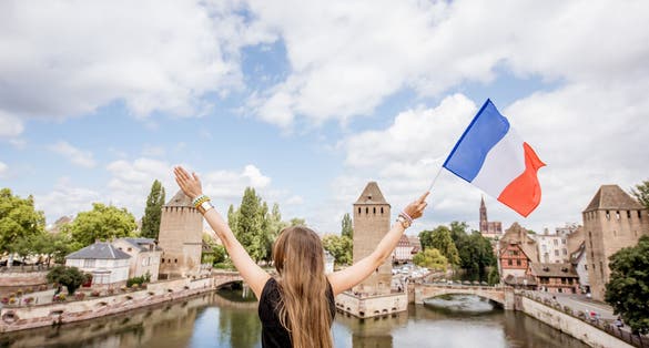 photo of young woman tourist sitting on Barrage Vauban with French flag on the beautiful landscape background with river and towers in the old town of Strasbourg city, France.