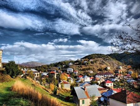 A peaceful village in North Macedonia, surrounded by hills, colorful houses, and a bright blue sky with clouds..png