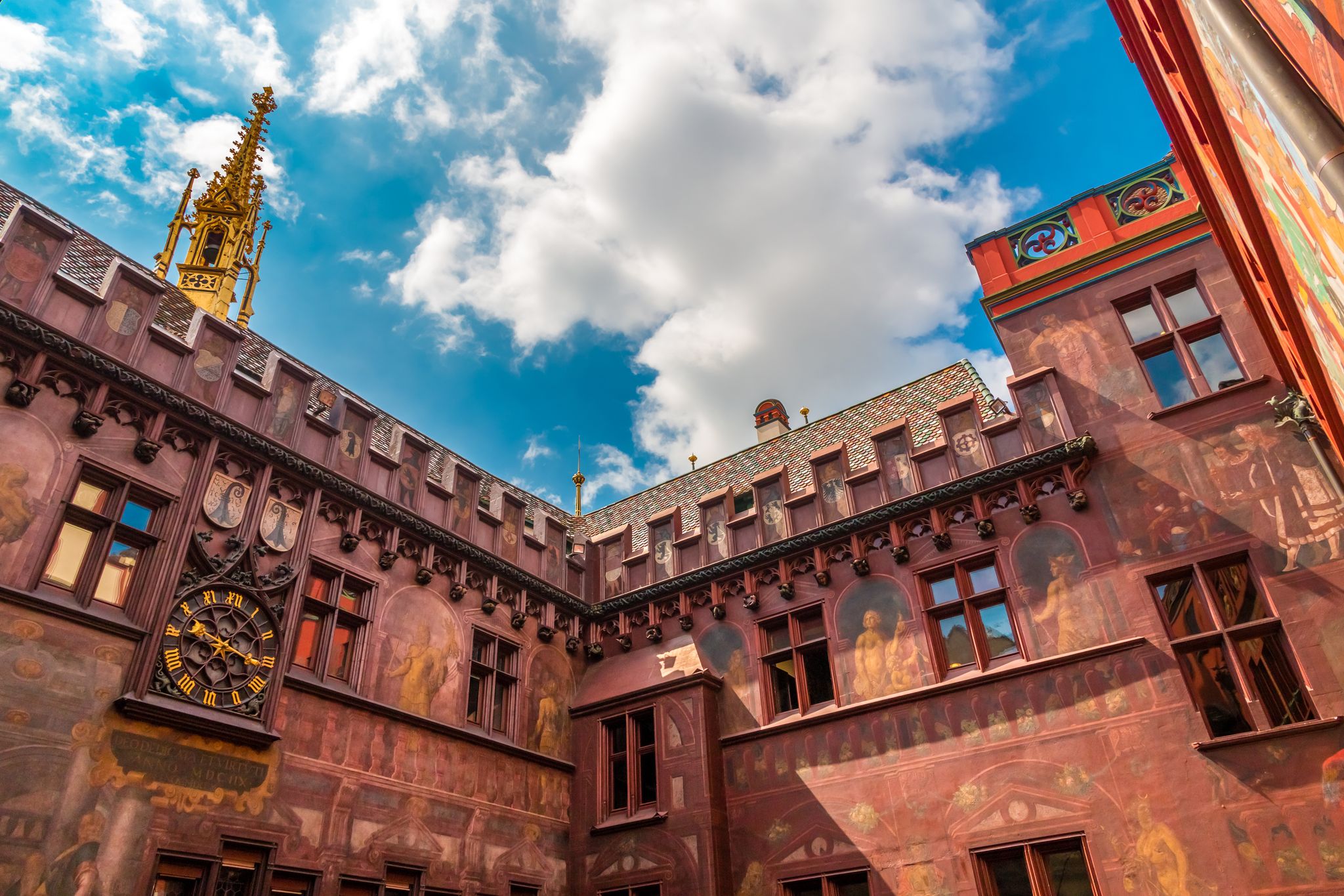 photo of great low-angle view of the inner courtyard of the Town Hall in Basel. It is characteristic for its colorful roof, red façade, frescoes, reliefs, crenellations, the wall clock and the golden turret in Basel, Switzerland.