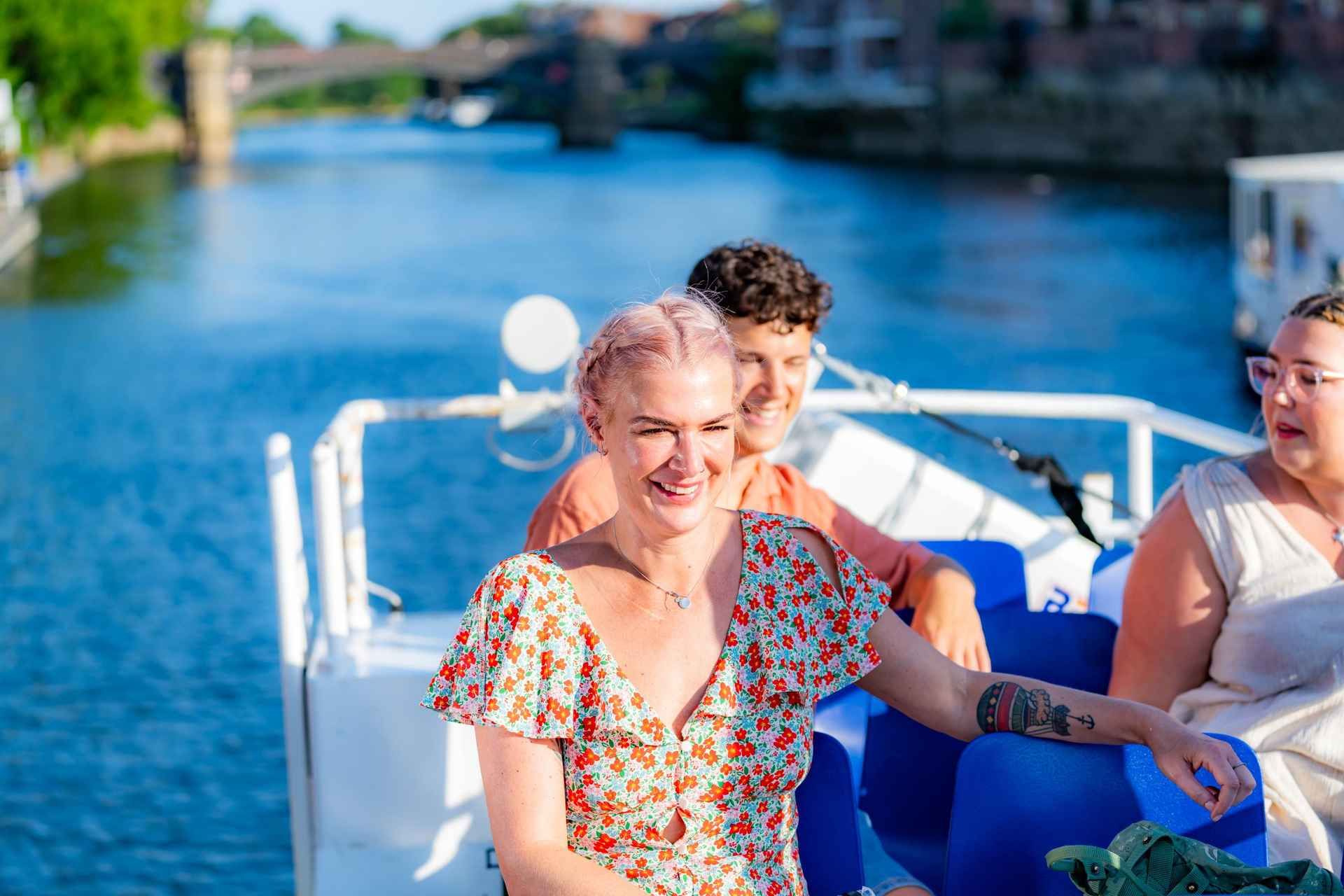 A smiling woman with braided hair enjoys a sunny boat ride with friends on the River Ouse, with a bridge in the background..jpg