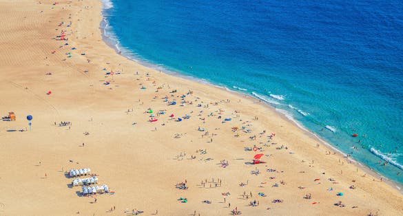 Top aerial view of sandy beach with people tourists sunbathing and Atlantic Ocean azure blue water, Praia da Nazare town coastline with waves, Leiria District, Oeste region, Portugal