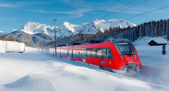 Photo of local train traveling by a forest in a valley covered by deep snow on a sunny winter day and Karwendel Mountain dominating the background under blue sky near Garmisch-Partenkirchen.
