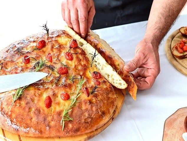 A person cuts a large, golden-brown focaccia bread topped with cherry tomatoes and rosemary on a wooden board..jpg