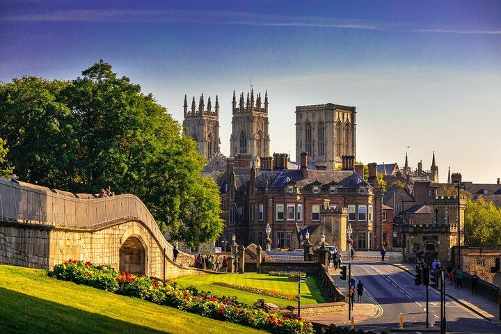 A sunny view of York Minster is surrounded by historic buildings, lush greenery, and a stone bridge in the foreground..jpg