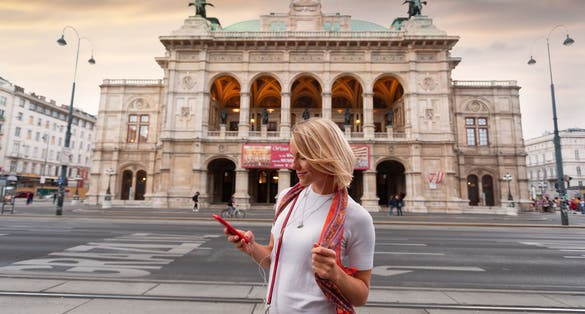 Photo of young beautiful smiling woman listens to music with headphones in front of Vienna State Opera at sunset, Austria.