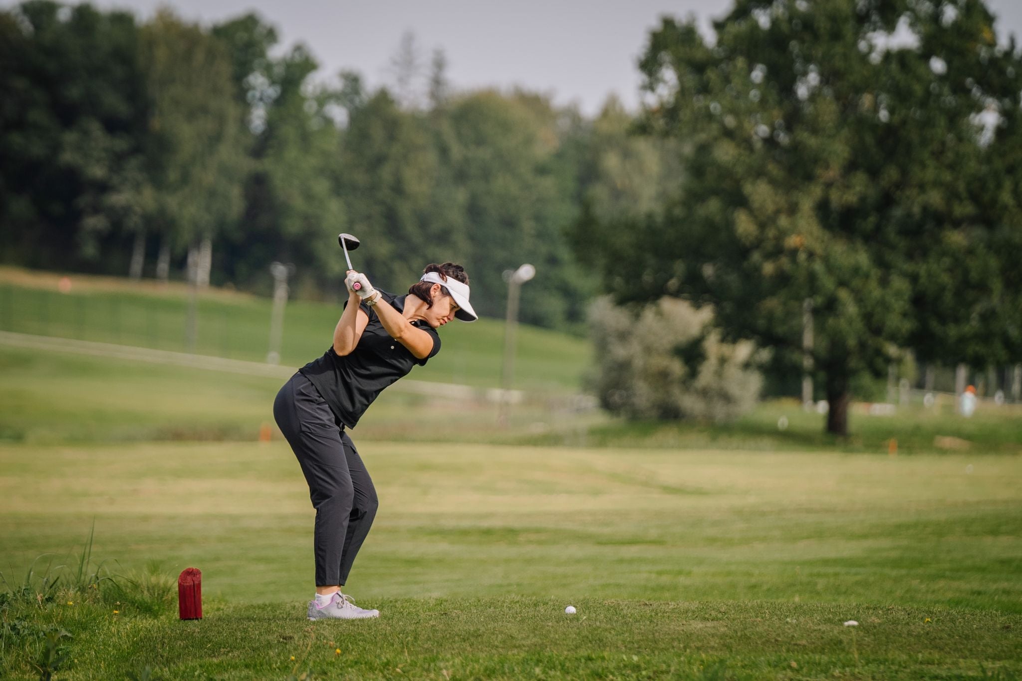 Sigulda, Latvia -  Woman golfer in black attire taking a swing on a green golf course, with trees and a red marker in the background.