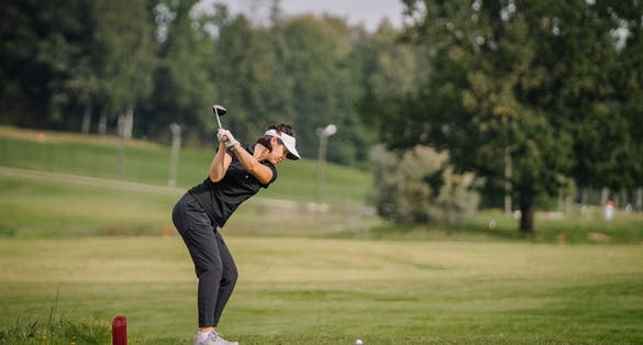 Sigulda, Latvia -  Woman golfer in black attire taking a swing on a green golf course, with trees and a red marker in the background.