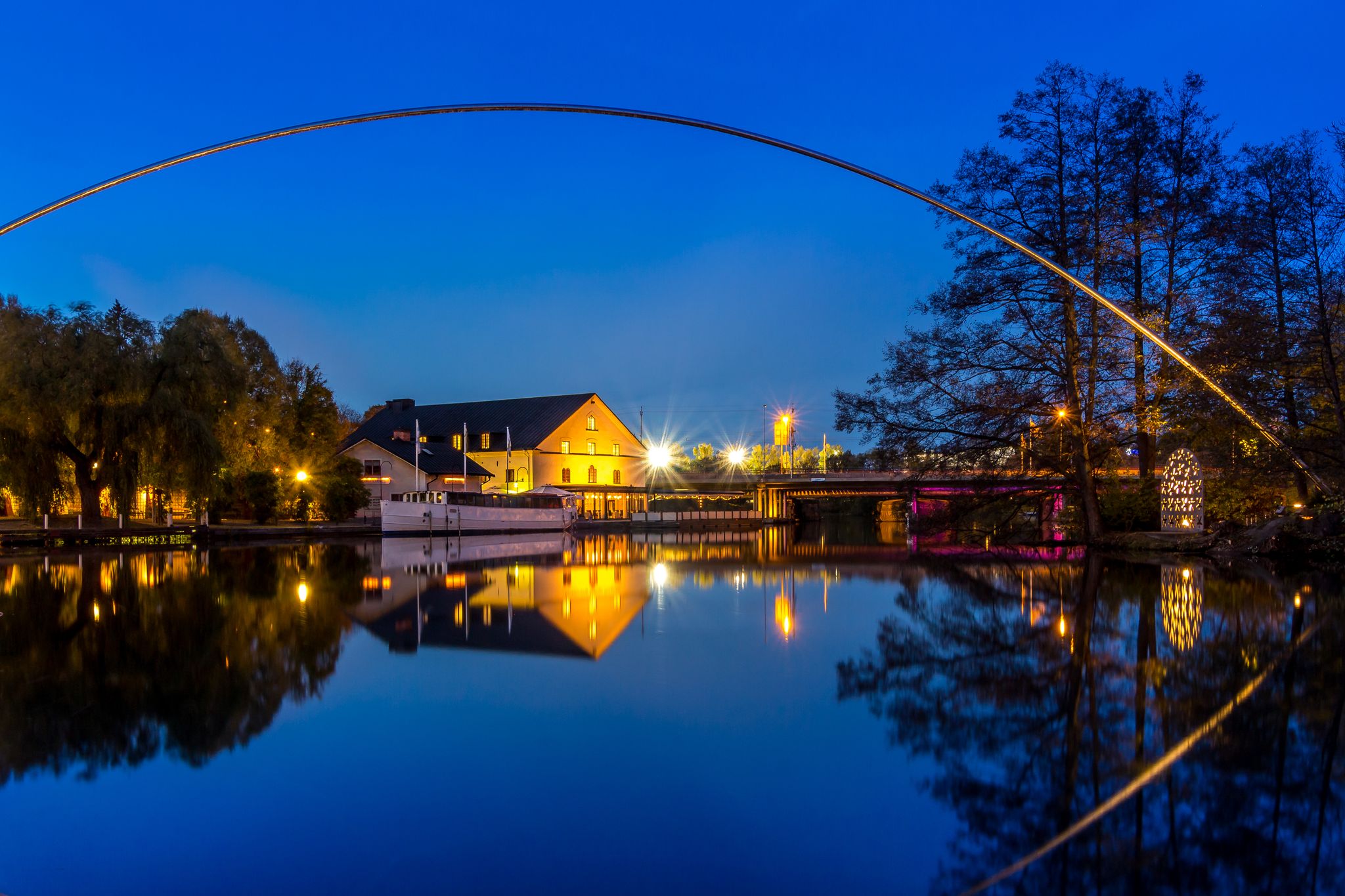 photo of Stangan or Kinda kanal at twilight time. Yellow lights shine on the Scandinavia building contrast with the blue sky. The metal curve structure reflects on the still water surface. Linköping, Sweden.