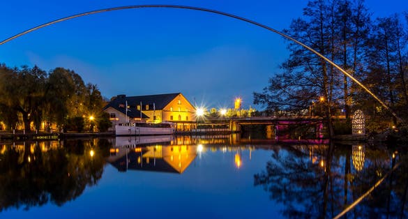 photo of Stangan or Kinda kanal at twilight time. Yellow lights shine on the Scandinavia building contrast with the blue sky. The metal curve structure reflects on the still water surface. Linköping, Sweden.
