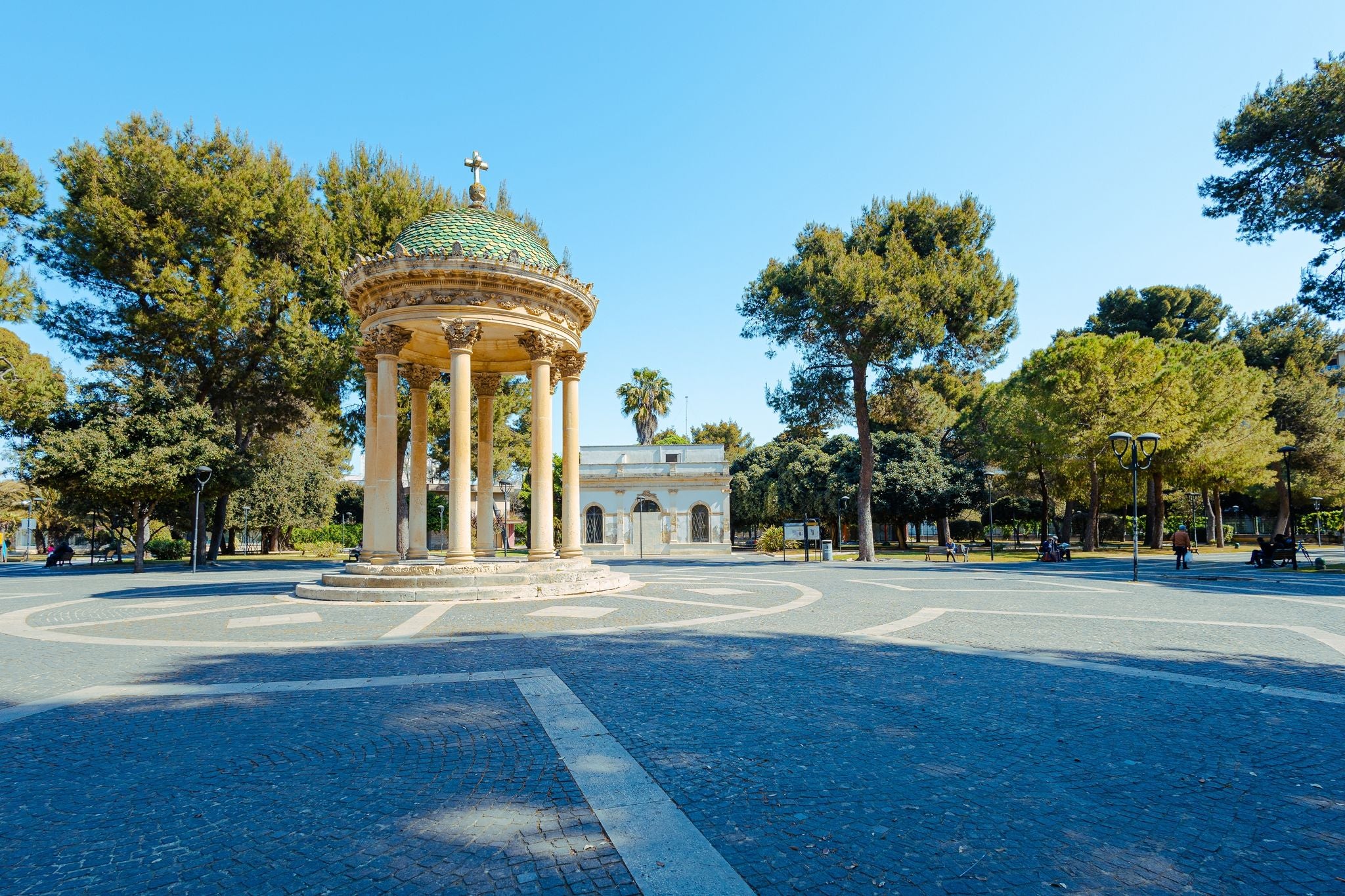 Central part of a big park in lecce, with round monument with pillars standing in the middle. Green trees and clear blue skies.