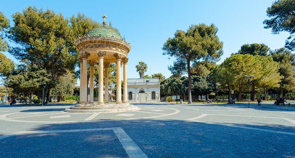 Central part of a big park in lecce, with round monument with pillars standing in the middle. Green trees and clear blue skies.