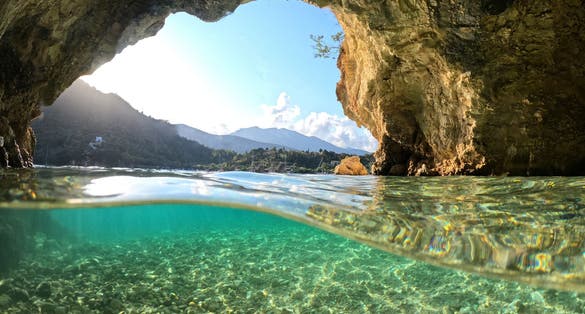 Photo of small white rock cave with crystal clear emerald sea and chapel of Agios Nikolaos.