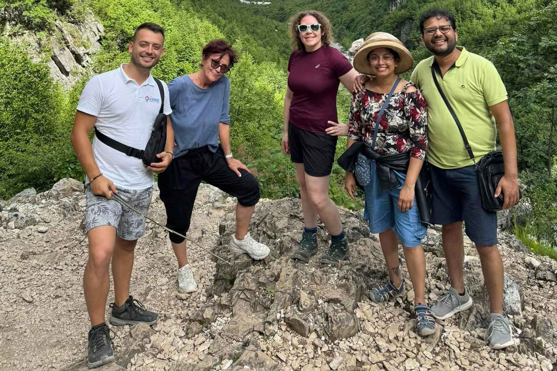 A group of five smiling hikers poses on a rocky trail surrounded by lush green mountains and trees in the Albanian Alps..jpg