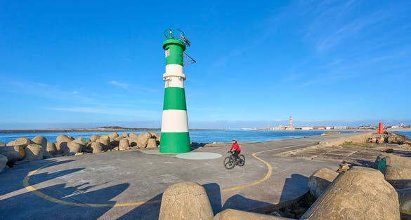 happy senior woman cycling below the navigation sign and ligthouse of Barra at the harbor entrance of Aveiro, Portugal