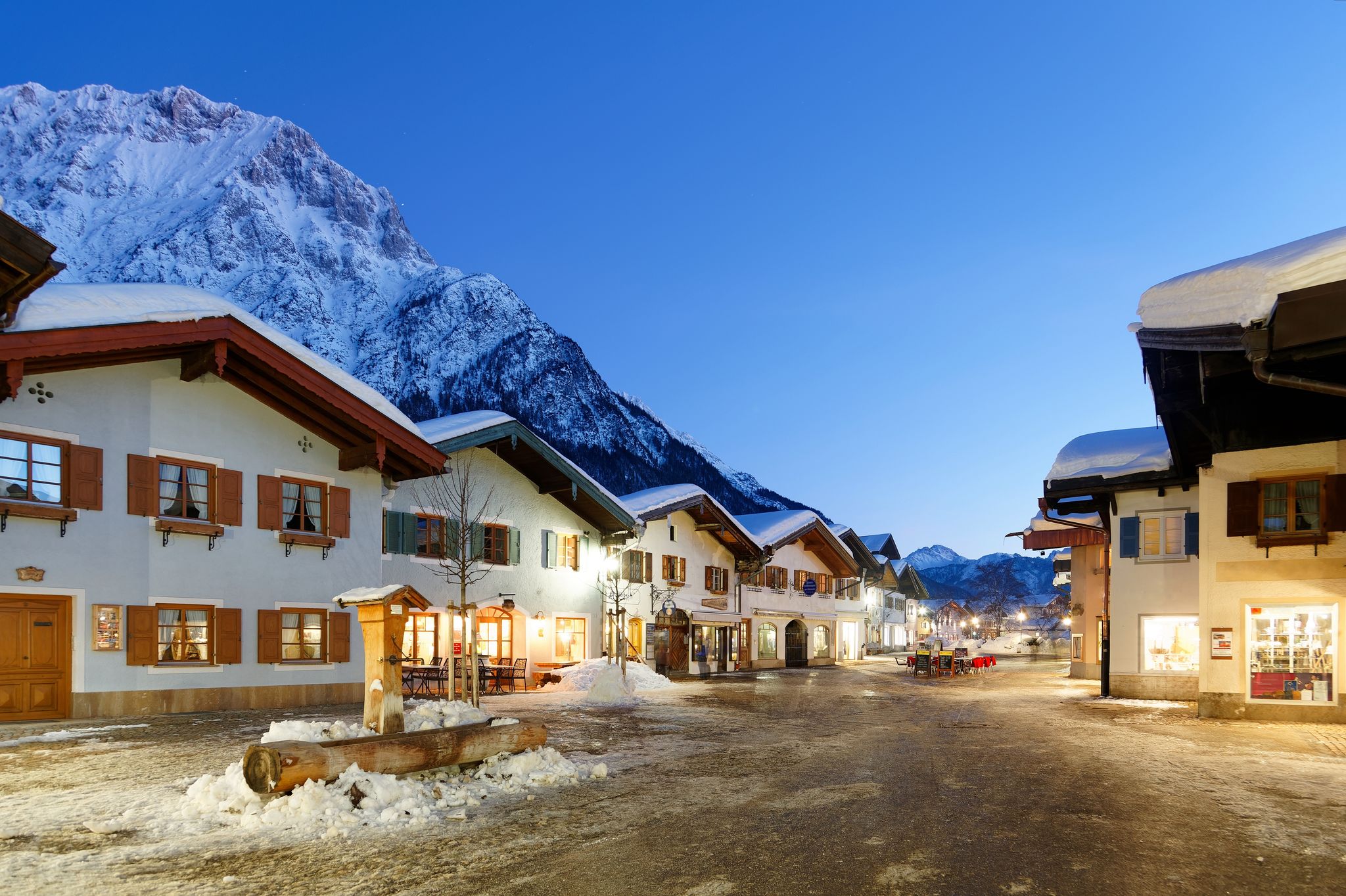 Photo of scenery of a street flanked by old chalet houses in blue twilight on a cold snowy winter evening in Mittenwald, Germany.
