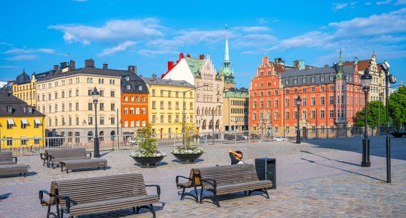 Benches line a cobblestone square in Stockholm bathed in sunlight, surrounded by historic colorful buildings under a clear blue sky. Sweden