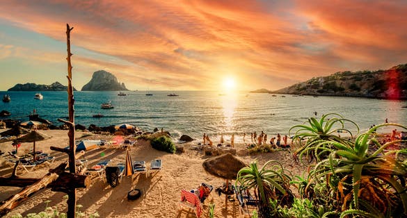 photo of Picturesque view of Cala d'Hort tropical Beach, people hangout in beautiful beach with Es Vedra rock view during magnificent vibrant sunset glowing sun. Balearic Islands, Spain.