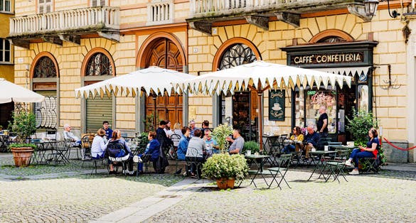 Turin, Italy. In Piazza della Consolata, under large umbrellas, outdoor tables of a cafeteria with some people sitting.