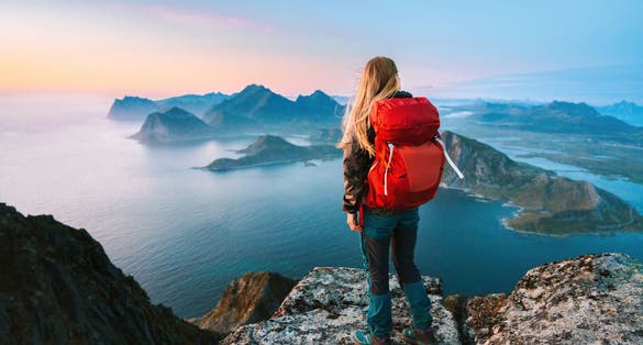 A female hiker with a red backpack stands on a rocky summit in Ryten, overlooking dramatic sea cliffs and islands in Norway’s Lofoten archipelago at sunset.