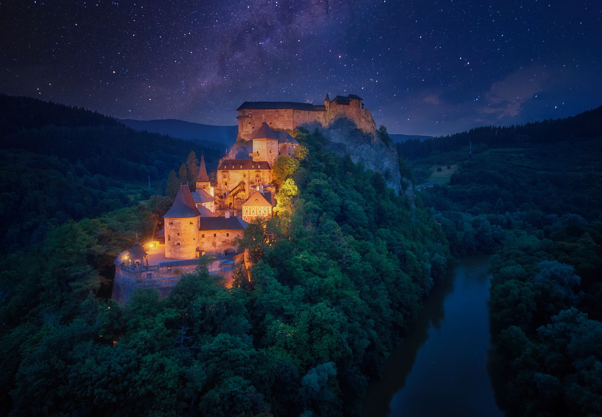 Night, aerial view of Orava Castle, fantasy castle situated on a high rock above Orava river, illuminated by orange lamps, surrounded by starry night over mountains. Castles of Slovakia.