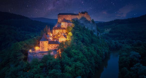 Night, aerial view of Orava Castle, fantasy castle situated on a high rock above Orava river, illuminated by orange lamps, surrounded by starry night over mountains. Castles of Slovakia.