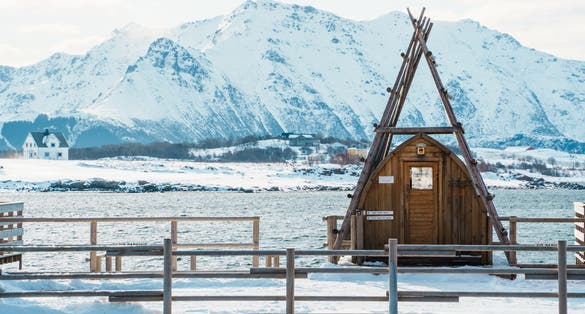 A small wooden sauna hut with a triangular roof sits by a snow-covered fjord near Leknes in Lofoten, Norway, with snowy mountain peaks in the background.