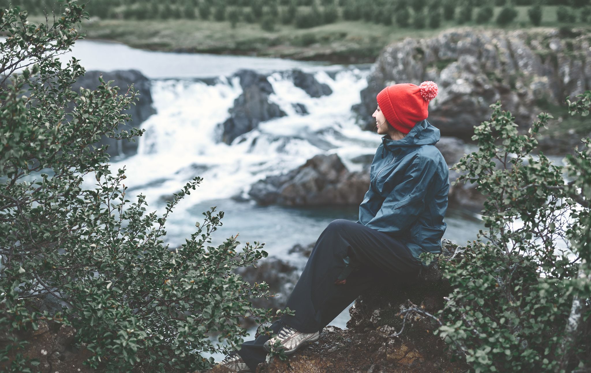 photo of a woman tourist in waterproof clothes and red hat sits on the Bank of the mountain river with small waterfall. Glanni waterfall, Iceland .