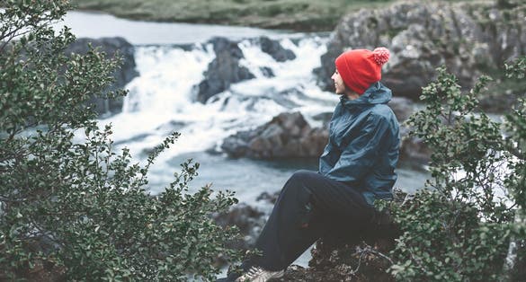 photo of a woman tourist in waterproof clothes and red hat sits on the Bank of the mountain river with small waterfall. Glanni waterfall, Iceland .