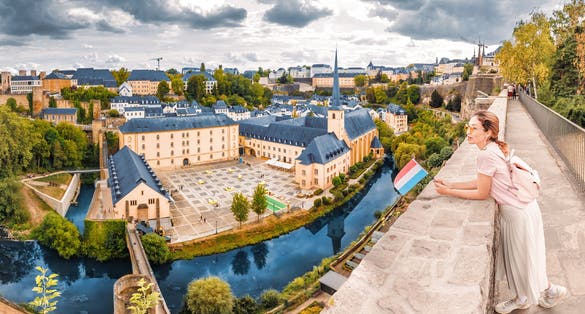 A happy Asian traveller girl holds the flag of Luxembourg and admires the great panoramic view of the Grund town from the viewing spot. Tourism, recreation and life in the country concept
