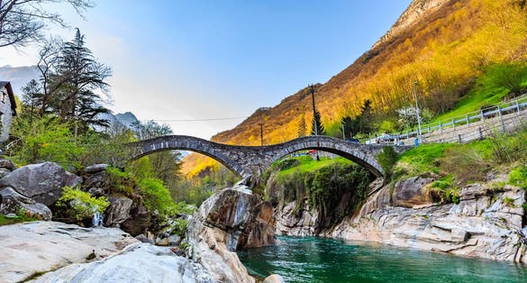 photo of two arches stone bridge soars over turquoise water stream of Valle Verzasca named Ponte dei Salti in canton of Ticino, Switzerland.