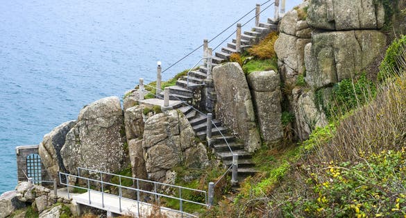 Photo of Steep steps descending between rocks and cliff with handrails beside the beautiful blue ocean at Minack Theatre near Porthcurno Beach, Cornwall, England.
