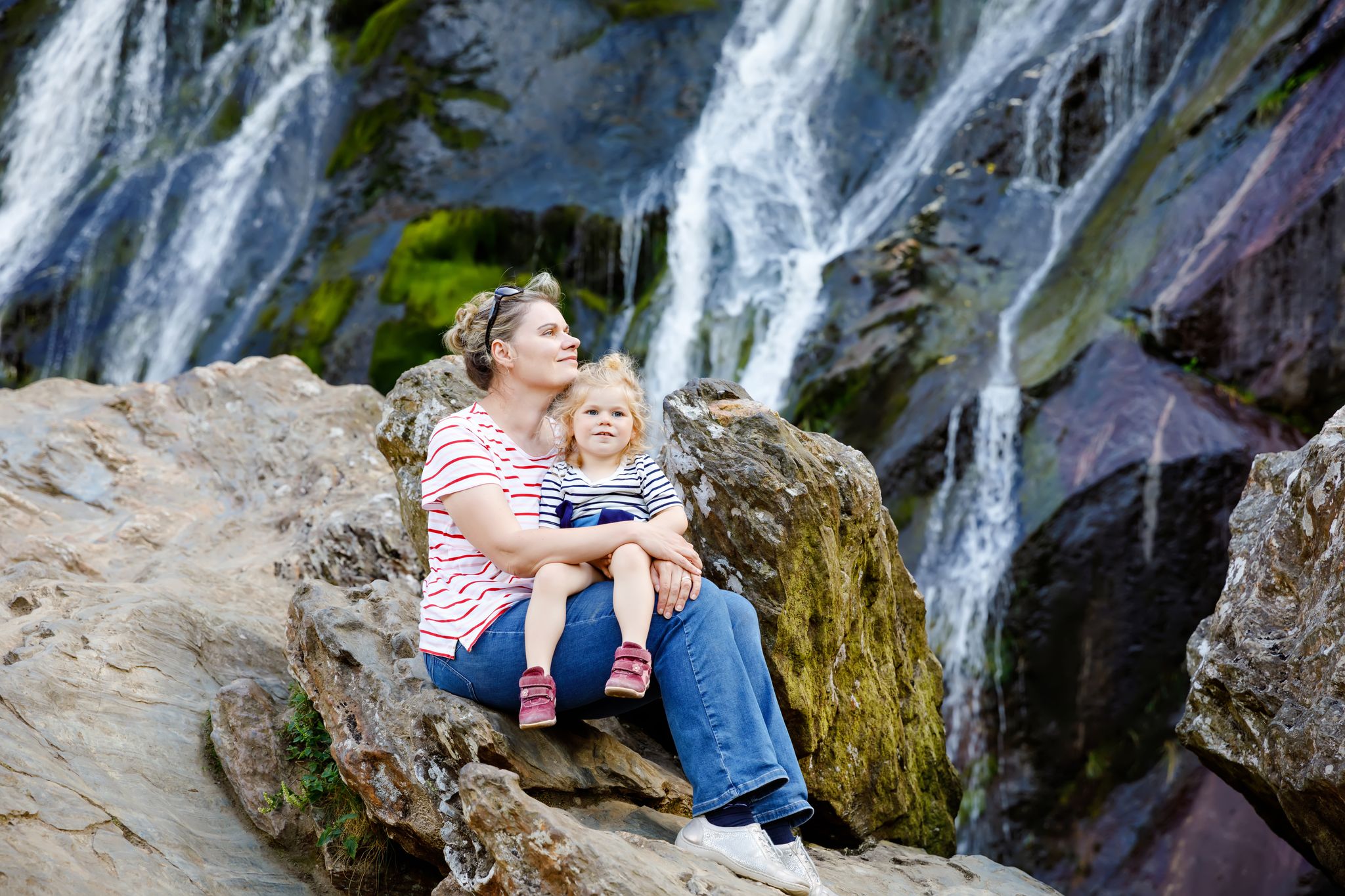 Cute toddler girl and mother sitting near water cascade of Powerscourt Waterfall, the highest waterfall in Ireland in co. Wicklow. 