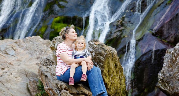 Cute toddler girl and mother sitting near water cascade of Powerscourt Waterfall, the highest waterfall in Ireland in co. Wicklow. 