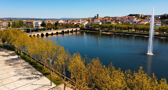 Photo of scenic view of Mirandela cityscape on banks of Tua River with landscaped embankment, modern water fountain and ancient stone arched bridge across river on sunny spring day, Portugal.