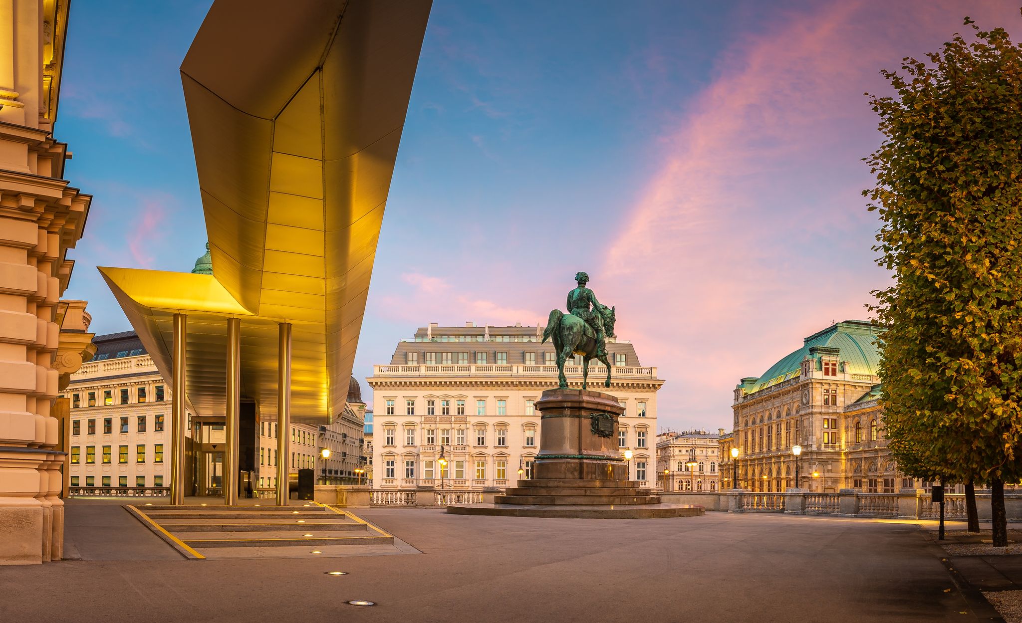Photo of square with statue in front an museum building Albertina in Vienna and historical hotel Sacher and Vienna state opera in the background during evening.