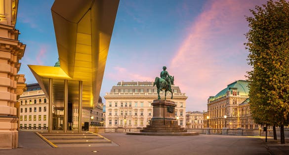Photo of square with statue in front an museum building Albertina in Vienna and historical hotel Sacher and Vienna state opera in the background during evening.