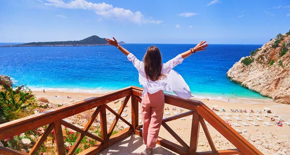 Photo of carefree traveler girl stands alone with open arms on stairs against backdrop of bay with the turquoise sea in Kas, Turkey.