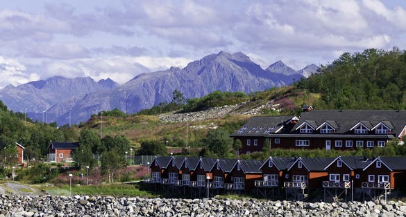 photo of view of Homes along shore protected by stone barrier, with mountains behind in Stokmarknes, Norway