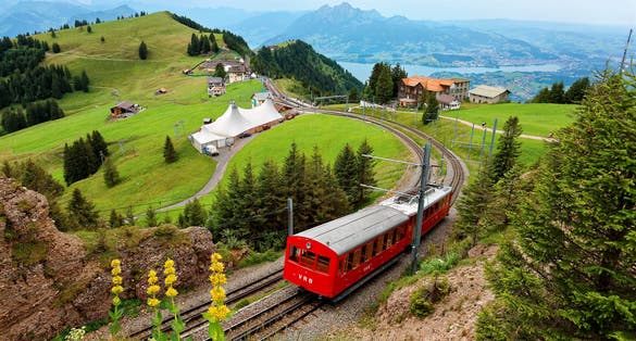 photo of a sightseeing train traveling on the cogwheel railway through green grassy meadows on Mt. Rigi, with rugged Pilatus peaks among majestic mountains in background on a cloudy summer day in Switzerland.