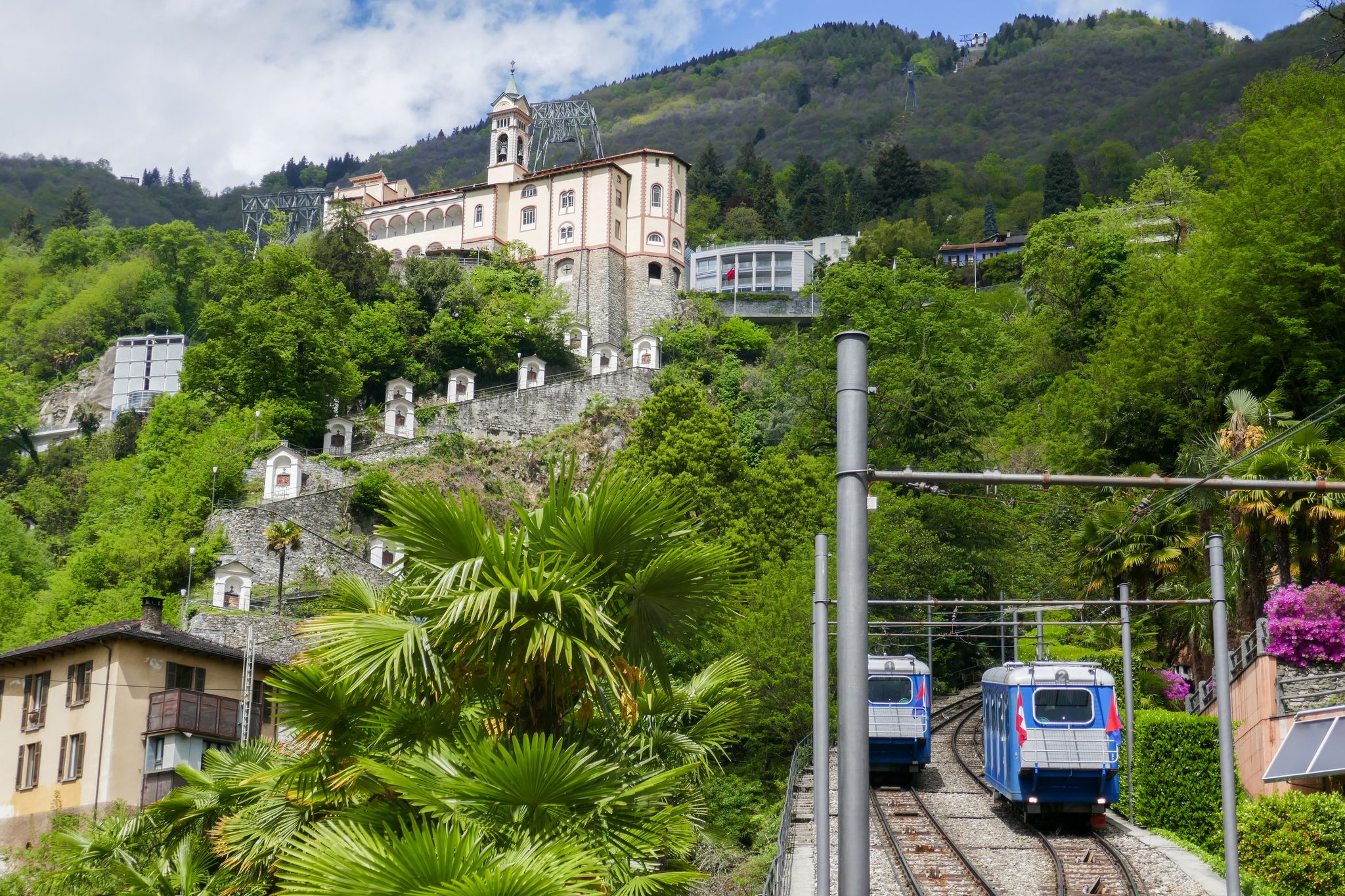 photo of two blue funicular trains on the rail tracks between Locarno and Orselina in Switzerland, with the famous Madonna del Sasso church and the "Kapellenweg" (Chapel Path) in the background in Locarno, Switzerland.