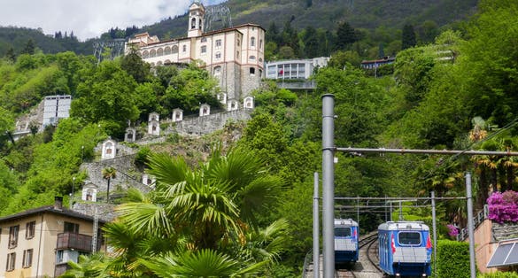 photo of two blue funicular trains on the rail tracks between Locarno and Orselina in Switzerland, with the famous Madonna del Sasso church and the "Kapellenweg" (Chapel Path) in the background in Locarno, Switzerland.