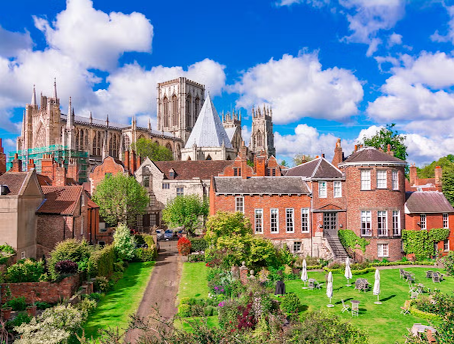 York Minster in York, England, with historic red-brick houses, lush green gardens, and a bright blue sky..png
