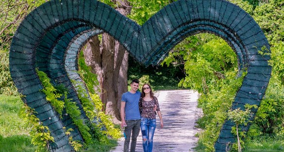 Young engaged couple walk under a green heart-shaped arch in the public park, Craiova, Romania