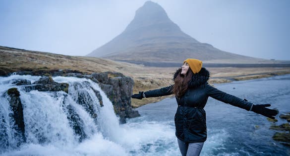 photo of Happy free beautiful girl dancing with open arms of happiness in the wind Iceland nature. In background on famous Kirkjufell Mountain with colorful, dramatic sky.Europe travel tourist enjoying wint