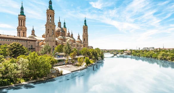 Photo of Scenery landscape with Basilica of Our Lady of Pillar. Blue sky reflects in clear water. Green trees along river.